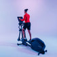 Man wearing red long-sleeve top and black shorts using black elliptical machine with digital display on gradient purple backg