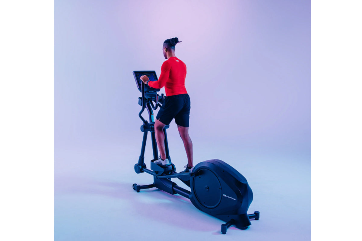 Man wearing red long-sleeve top and black shorts using black elliptical machine with digital display on gradient purple backg