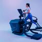 Woman using black Star Trac elliptical machine with digital display, wearing light blue t-shirt and blue leggings, against gr