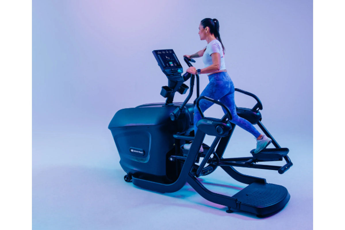 Woman using black Star Trac elliptical machine with digital display, wearing light blue t-shirt and blue leggings, against gr