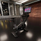 Black stationary exercise bike with digital screen mounted above handlebar, positioned in modern industrial studio setting wi