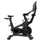 Black stationary exercise bike with tablet display showing pink interface, adjustable seat and handlebars, on white backgroun