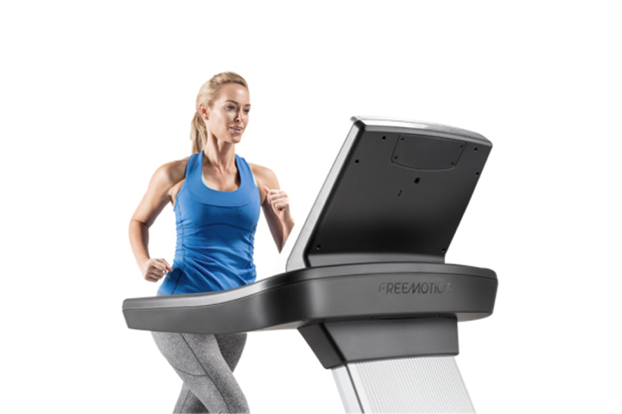 Woman in blue sports bra and gray leggings running on black Freemotion t10.9 treadmill with digital display against white bac