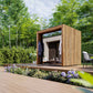 Modern wooden outdoor sauna with vertical slat design and glass doors in garden setting surrounded by green trees and floweri