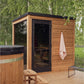 Cedar wood outdoor sauna with black roof and glass door, mounted on wooden deck with ivy-covered wall backdrop