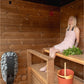 Woman wearing pink halter dress sitting on wooden bench inside sauna with heater and ladle visible