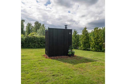 Black vertical wood-slat outdoor sauna with chimney in landscaped garden surrounded by green lawn and evergreen trees