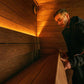 Man wearing black jacket inside wooden sauna with warm lighting, demonstrating interior bench seating