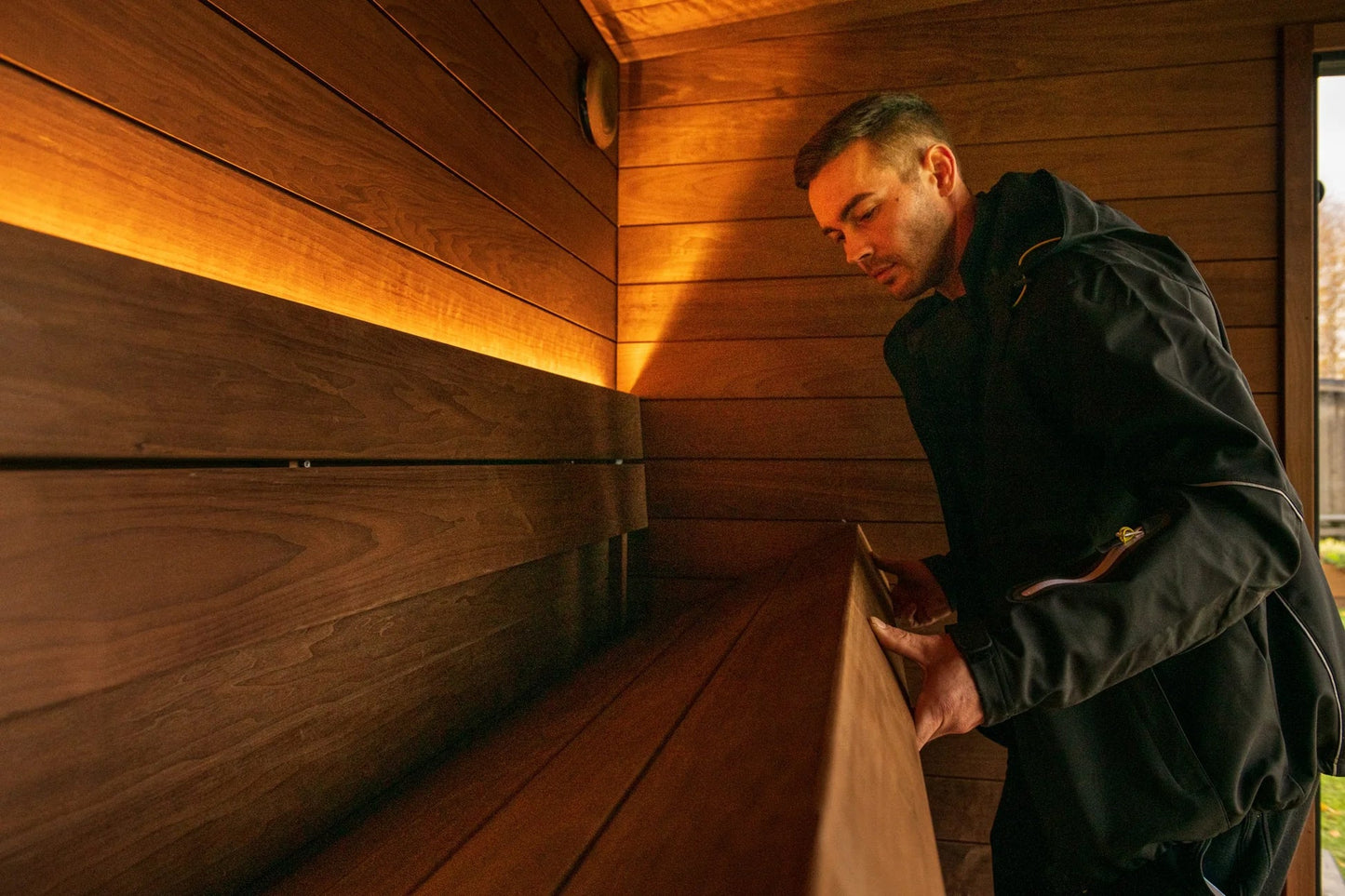 Man wearing black jacket inside wooden sauna with warm lighting, demonstrating interior bench seating