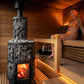 Woman in orange bikini sitting on wooden bench inside modern sauna with lit wood stove and warm ambient lighting