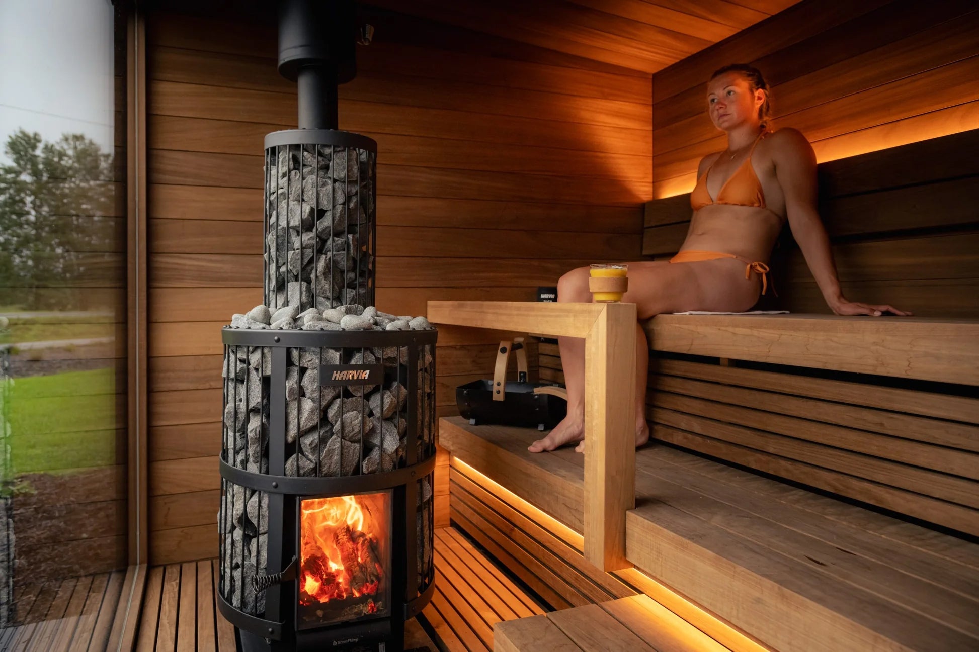 Woman in orange bikini sitting on wooden bench inside modern sauna with lit wood stove and warm ambient lighting
