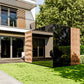 Modern wooden outdoor sauna with vertical slat design positioned on lawn beside contemporary architecture and green hedge wal