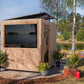 Modern wooden sauna cabin with large dark window, black roof trim, and entry door on lakeside setting surrounded by trees and