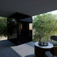Black outdoor sauna with glass doors on modern patio, surrounded by green trees and contemporary furniture
