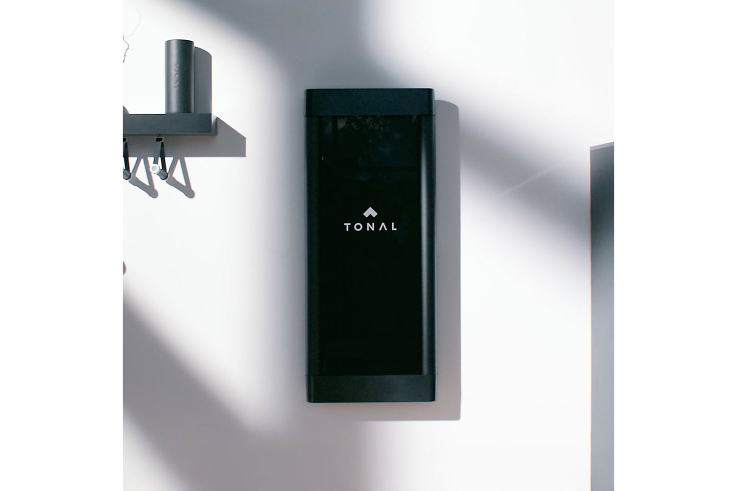 Black rectangular Tonal smart home gym display mounted on white wall with gray metal mounting bracket and cylindrical post