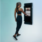 Woman wearing black sports bra and leggings interacting with wall-mounted black Tonal smart home gym displaying workout inter