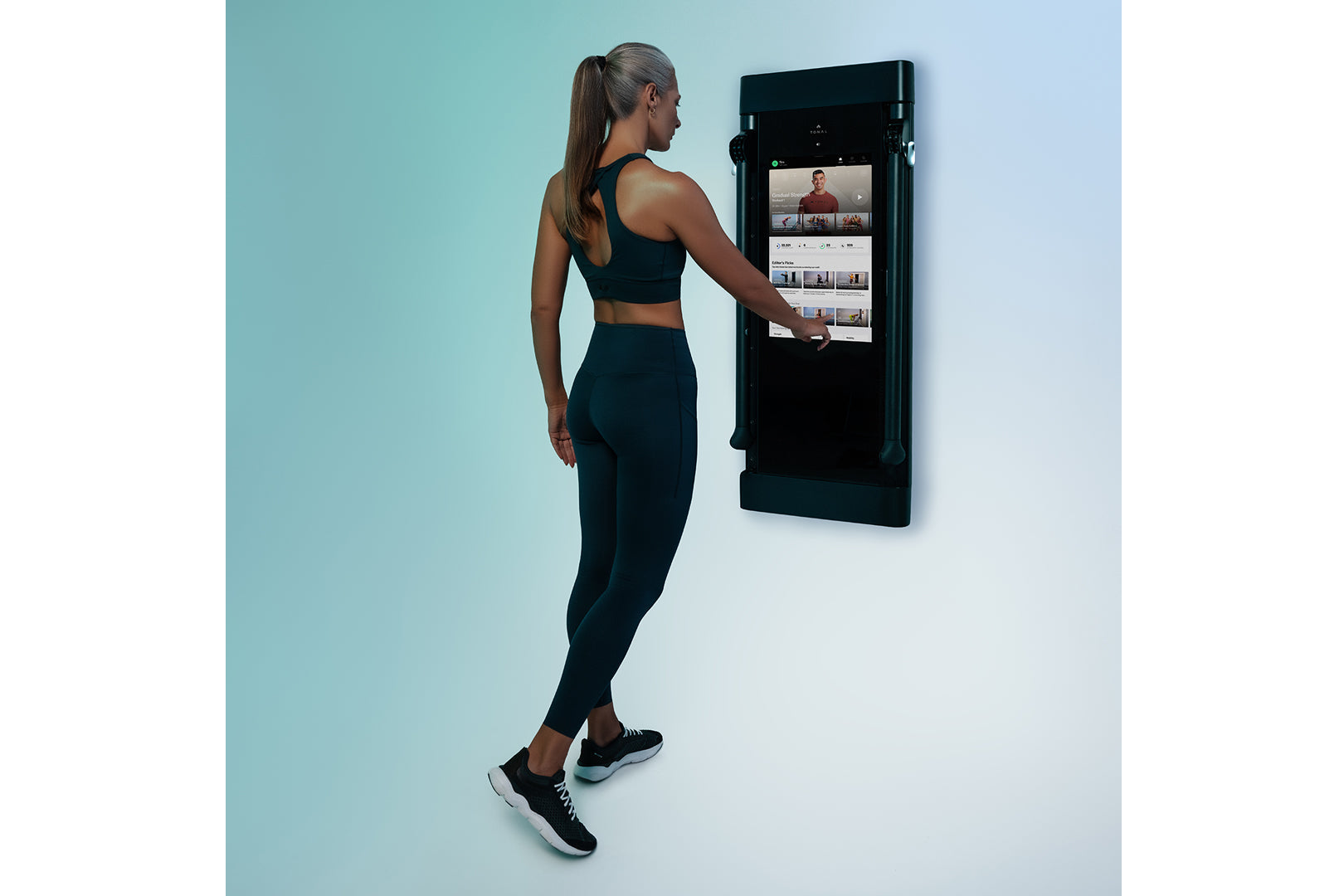 Woman wearing black sports bra and leggings interacting with wall-mounted black Tonal smart home gym displaying workout inter