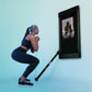 Woman doing a squat exercise with resistance band connected to black wall-mounted smart fitness mirror displaying workout vid