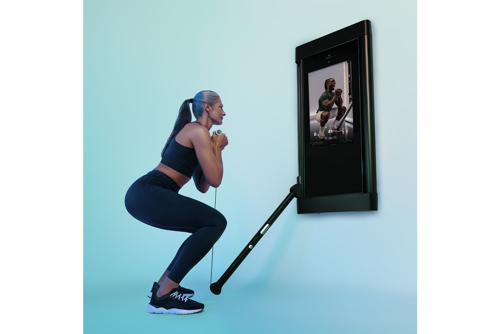Woman doing a squat exercise with resistance band connected to black wall-mounted smart fitness mirror displaying workout vid