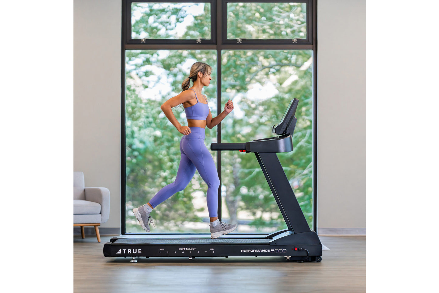 Woman in purple sports bra and leggings running on black TRUE Performance 8000 treadmill in modern room with large windows ov