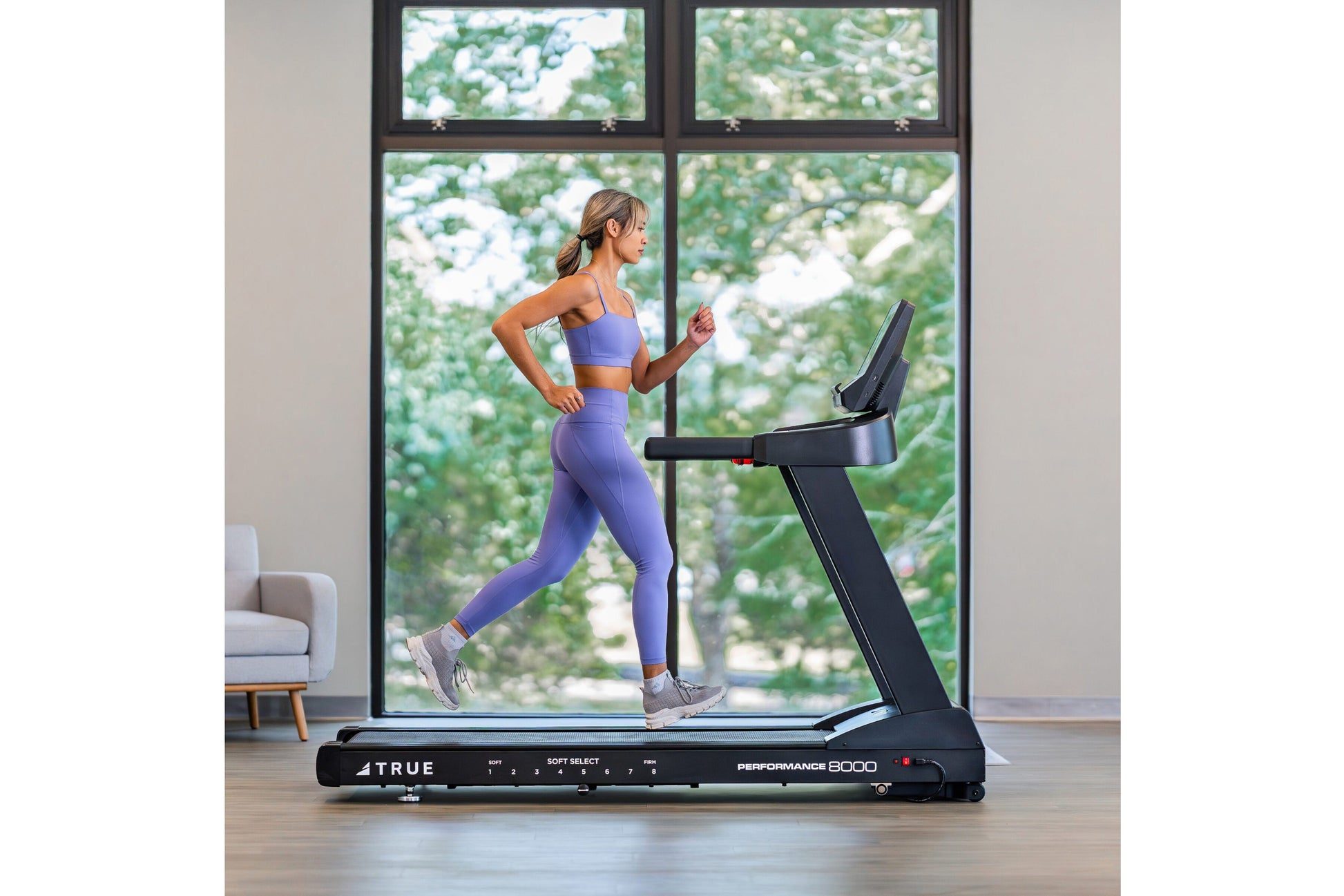 Woman in purple sports bra and leggings running on black TRUE Performance 8000 treadmill in modern room with large windows ov