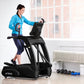 Woman in blue jacket and navy leggings using black TRUE elliptical machine in bright gym setting with dumbbells nearby