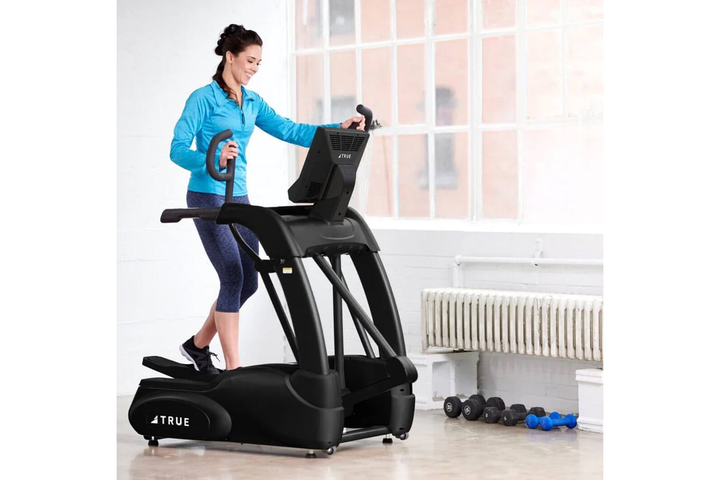 Woman in blue jacket and navy leggings using black TRUE elliptical machine in bright gym setting with dumbbells nearby