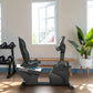 Black recumbent exercise bike with blue accents and digital display in modern gym setting with dumbbells and windows