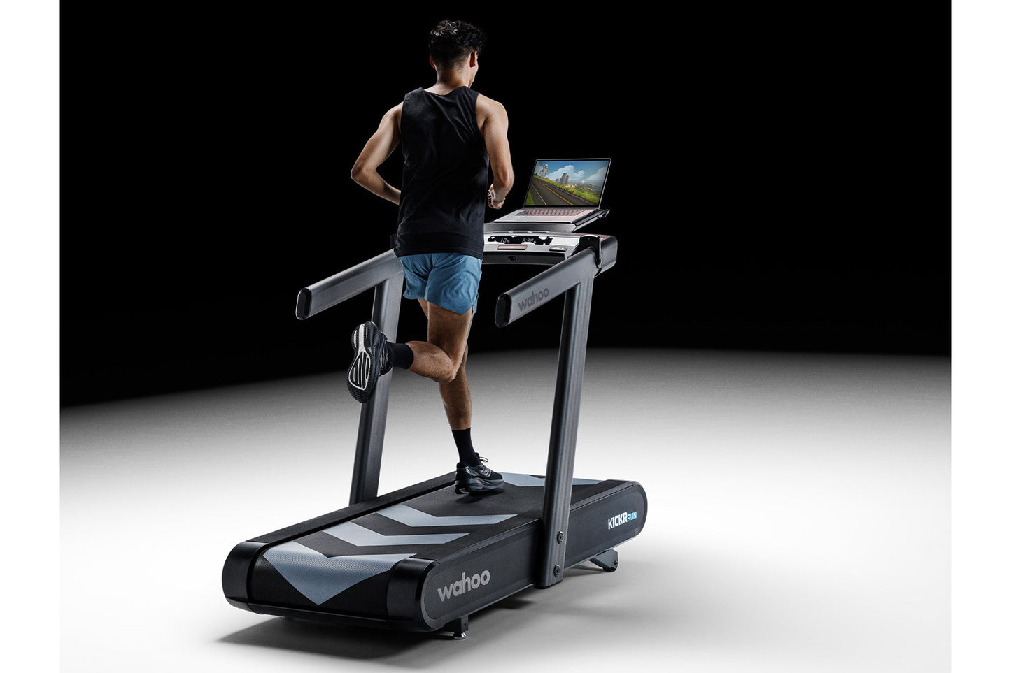 Man running on black and gray Wahoo KICKR RUN smart treadmill with laptop displaying scenic route on desk, worn by model on b