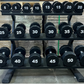 Black 12-sided urethane dumbbells arranged on tiered metal rack, labeled 5 to 50 pounds, in gym setting