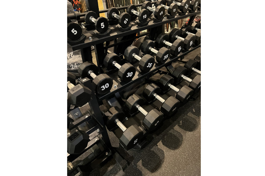 Black hexagonal urethane dumbbells arranged on tiered metal rack in gym setting, weights ranging from 5 to 50 pounds labeled 