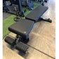 Black and blue adjustable weight bench with leg extension attachment and foot rollers, displayed in gym setting on concrete f