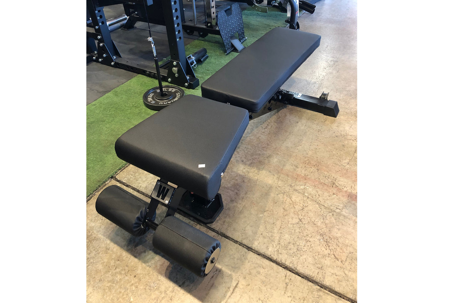 Black and blue adjustable weight bench with leg extension attachment and foot rollers, displayed in gym setting on concrete f