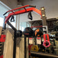 Red and black metal power rack with pull-up bar, weight plates, and dumbbells displayed in gym setting