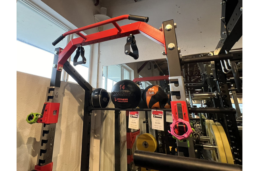 Red and black metal power rack with pull-up bar, weight plates, and dumbbells displayed in gym setting