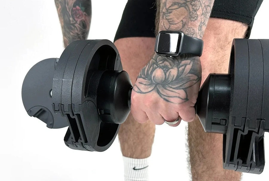 Man holding pair of black adjustable dumbbells with square buckle straps, wearing black shorts and white socks on white backg
