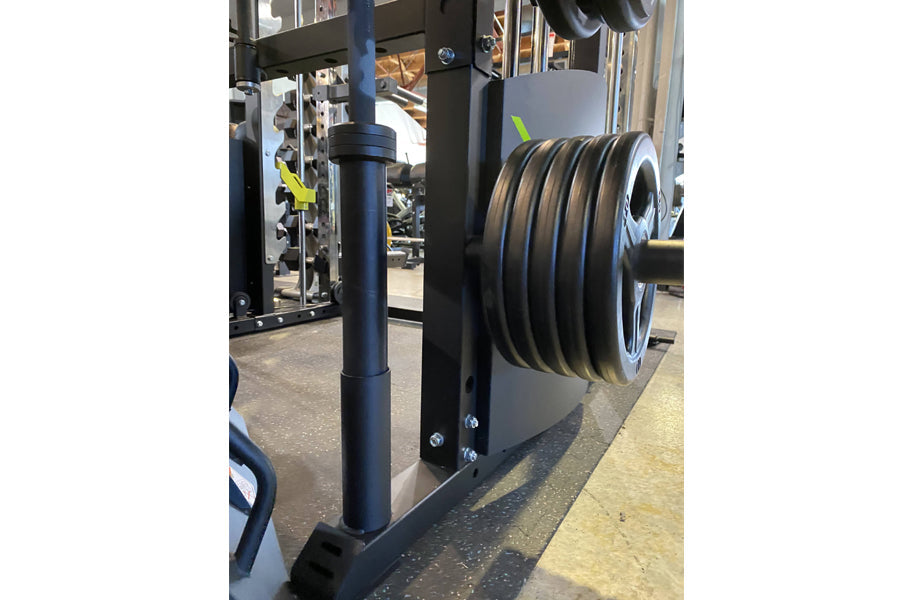 Black steel power rack with stacked weight plates loaded on right side, gym equipment in background