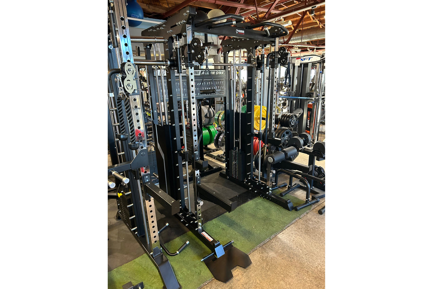Black multi-functional power rack with cable pulleys, weight plates, and dumbbells on green and black gym flooring in warehou