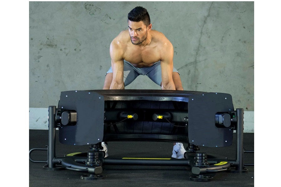 Shirtless man in blue shorts performing tire flip exercise on black Warrior Tire Flip 180 equipment against concrete wall
