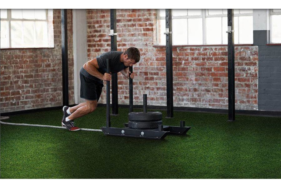 Man performing push exercise on black sled system with tire, on green turf in industrial gym space
