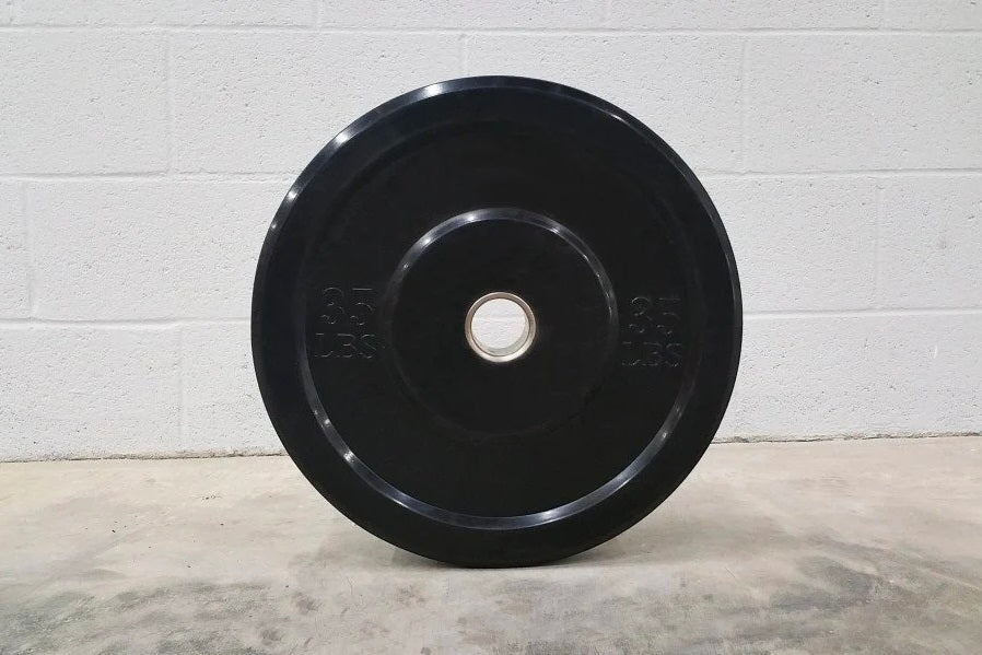 Black rubber bumper plate with 25kg weight markings and metal center sleeve, displayed on concrete floor against white brick 