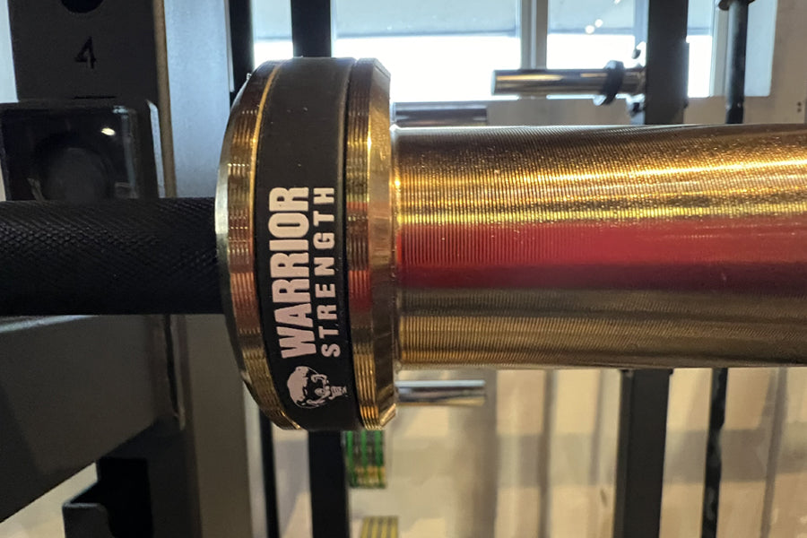 Warrior Strength black cerakote and gold titanium barbell with red and gold striped sleeve, displayed in gym setting
