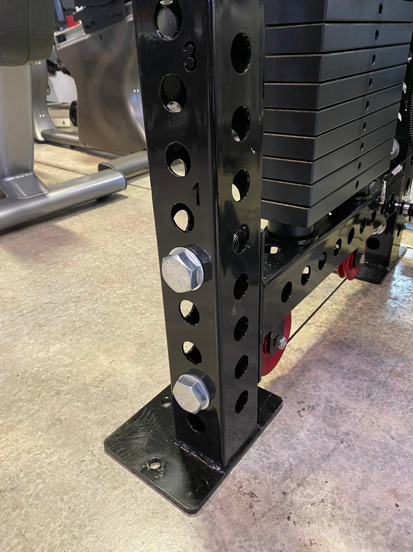 Black steel weight stack machine with multiple circular holes and chrome adjustment pins, displayed in gym setting