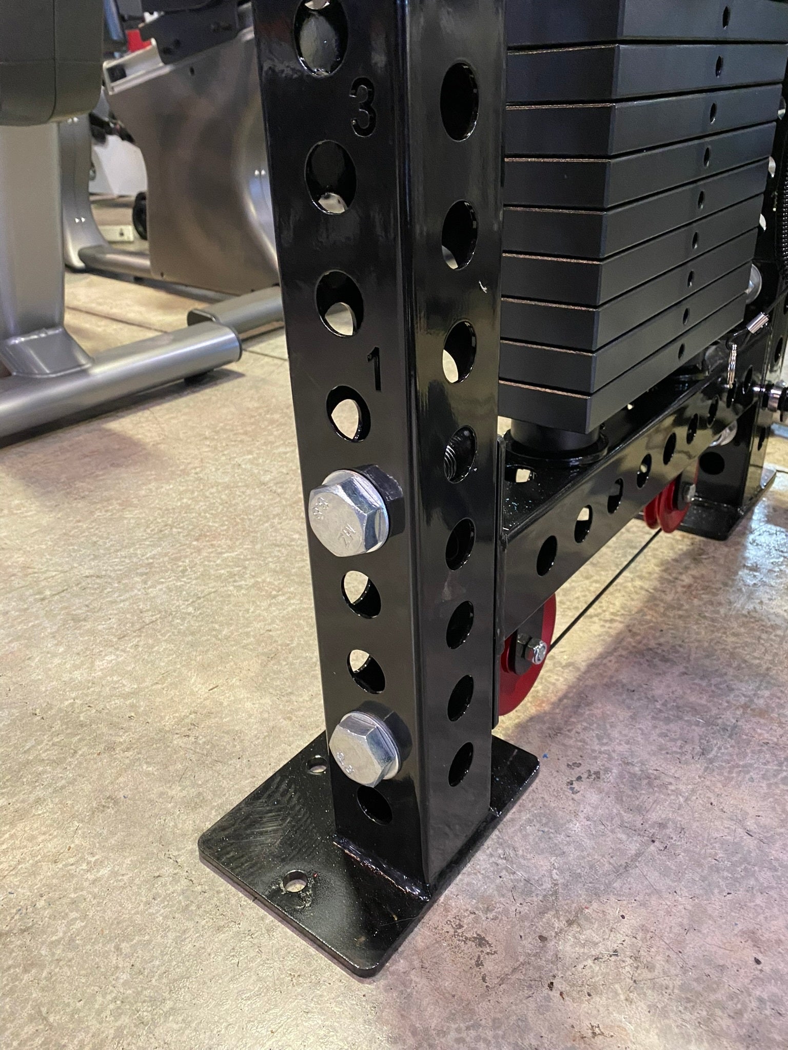 Black steel weight stack machine with multiple circular holes and chrome adjustment pins, displayed in gym setting