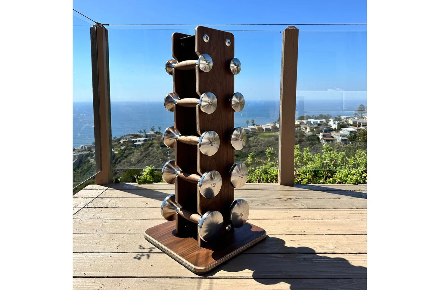 Dark wood dumbbell tower with stainless steel handles holding five pairs of weights, displayed on wooden deck with ocean view