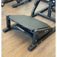 Black V-squat machine with gray cushioned platform, adjustable backrest, and metal frame on wooden gym floor