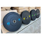 Row of black rubber bumper plates with weight markings in blue and yellow, displayed on gray floor with cardboard boxes in ba