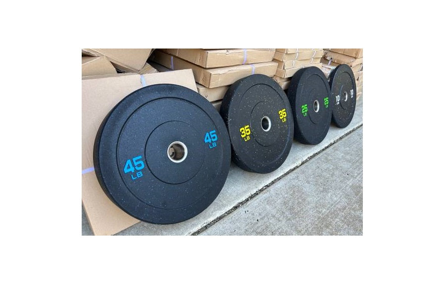 Row of black rubber bumper plates with weight markings in blue and yellow, displayed on gray floor with cardboard boxes in ba