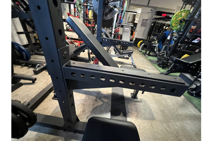 Black steel squat rack with angled uprights and perforated safety bars, shown in warehouse setting
