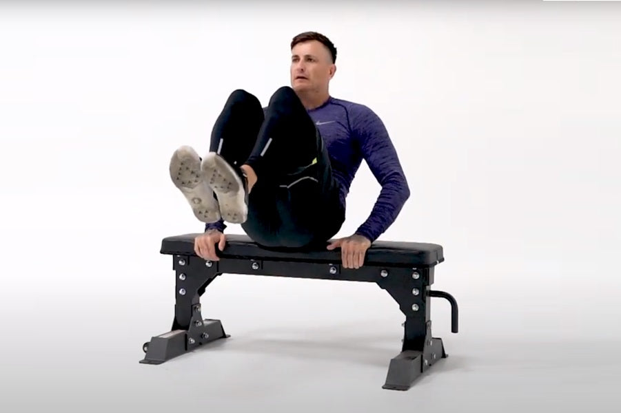 Man wearing blue long-sleeve shirt and black pants sitting on black Warrior flat bench with feet up, white background